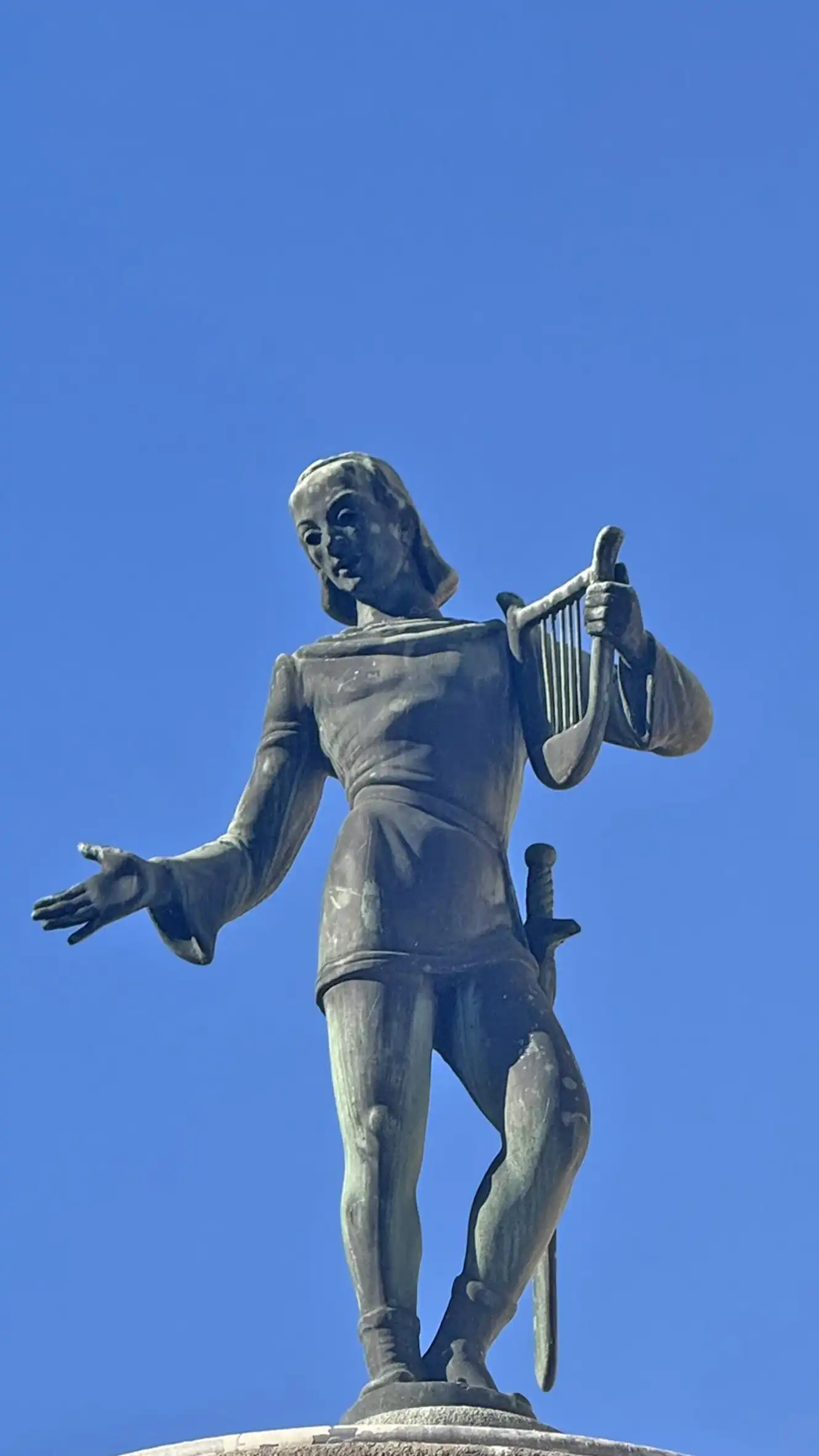 The medieval poet is standing on top of the fountain and holding a harp.