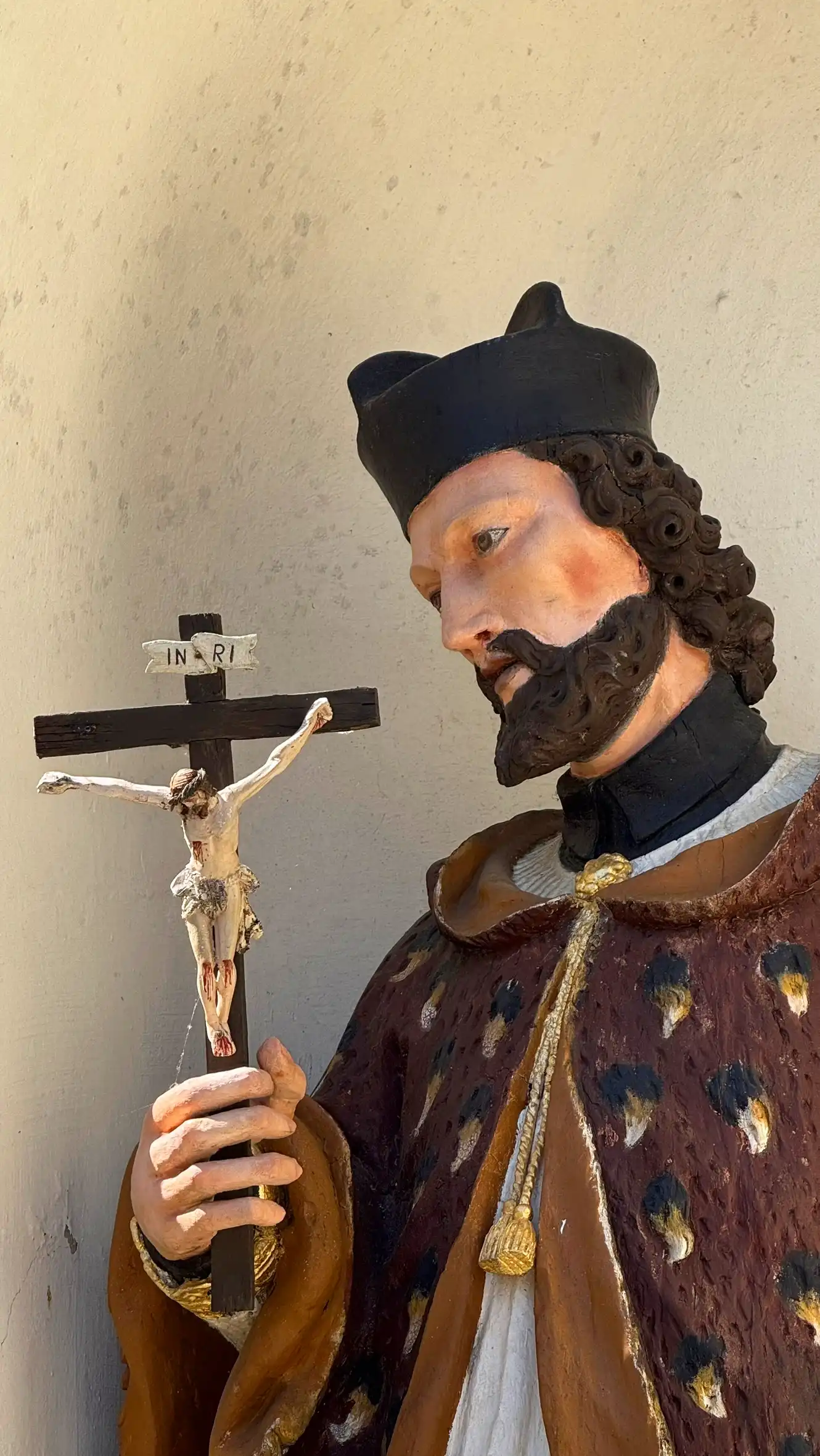 St John of Nepomuk is shown with curly hair and a dark beard. He is wearing a black, circular biretta.