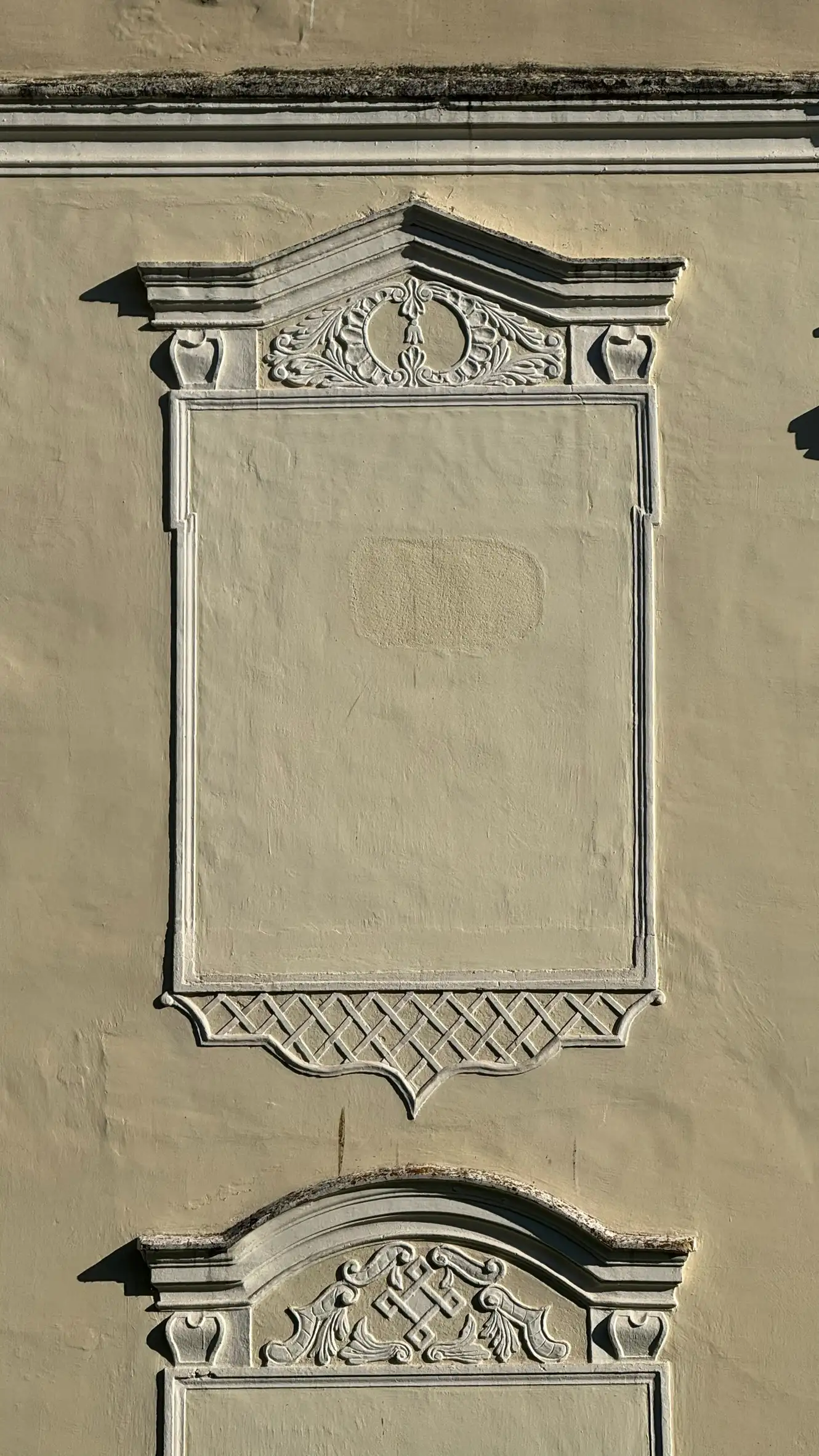 Nahaufnahme eines rechteckigen Blindfensters mit skulptiertem Rahmen und Giebel; Ornamentik ober- und unterhalb des Blindfensters