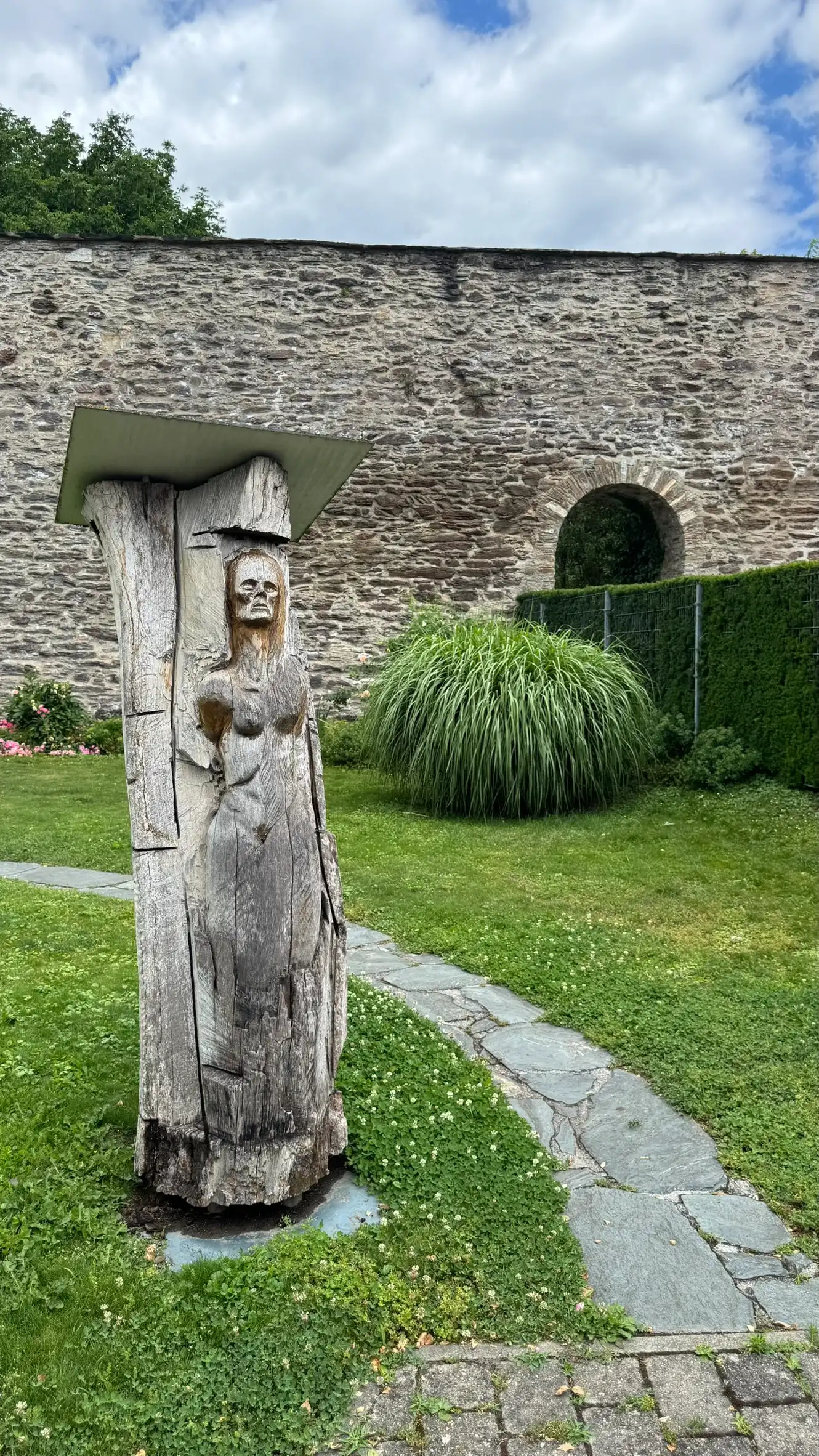 Vordergrund: Skulptur aus einem geschnitzten Baumstamm und geschwungener Weg; Hintergrund: NW Stadtmauer.